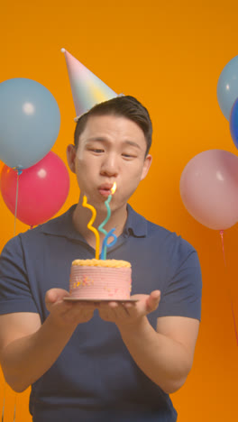 Retrato-De-Estudio-De-Vídeo-Vertical-De-Un-Hombre-Vestido-Con-Sombrero-De-Fiesta-Celebrando-Un-Cumpleaños-Soplando-Velas-En-El-Pastel